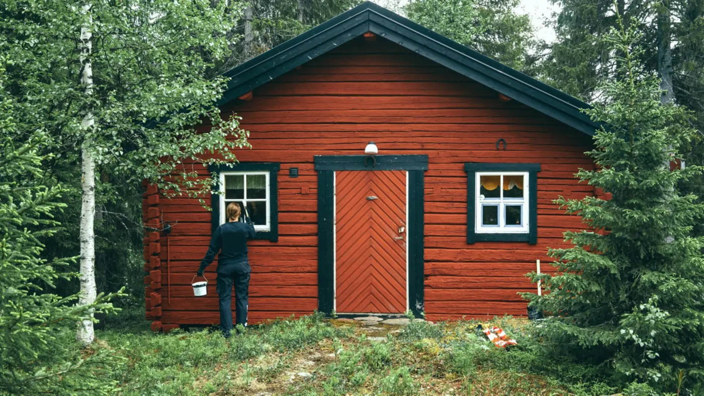 Målare med färghink framför en falurödmålad timmerstuga